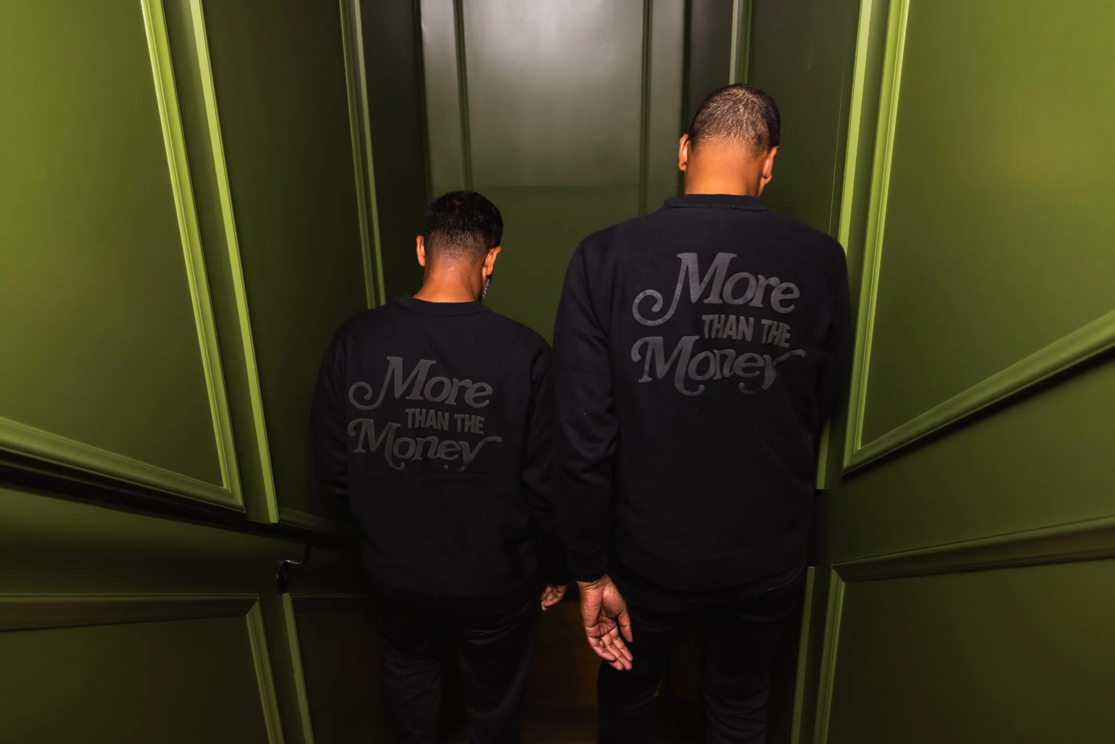 Two members of the MUUS crew standing together in matching "More Than the Money" knits at the end of a green corridor
